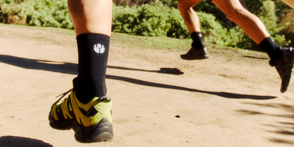 Close-up of a runner's black Fractel mens running socks with a white logo on a sunny dirt trail