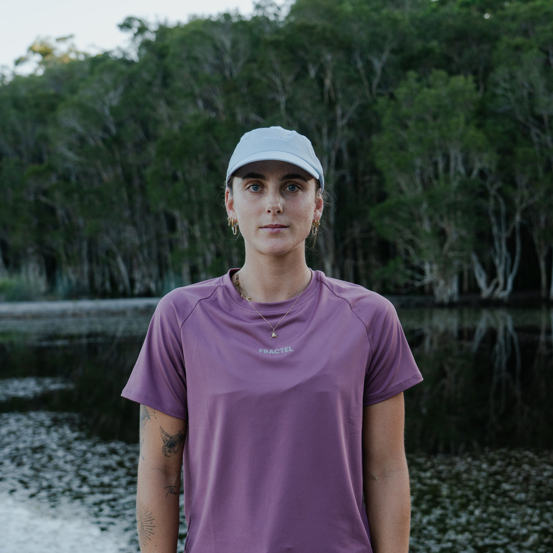 Female wearing a purple shirt and white cap standing in a natural setting with trees and water.