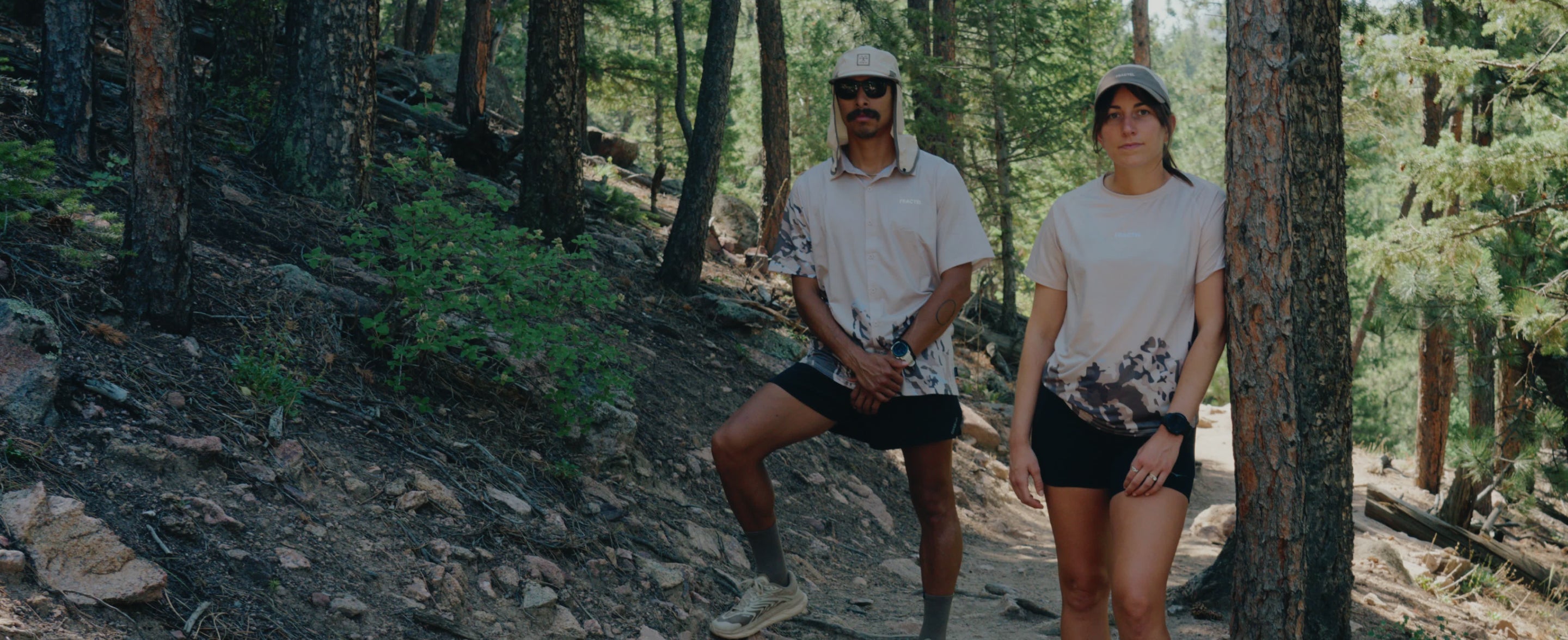A man and a woman standing on a forest trail wearing matching light beige short-sleeve t-shirts with a dark mountainous print and black shorts.