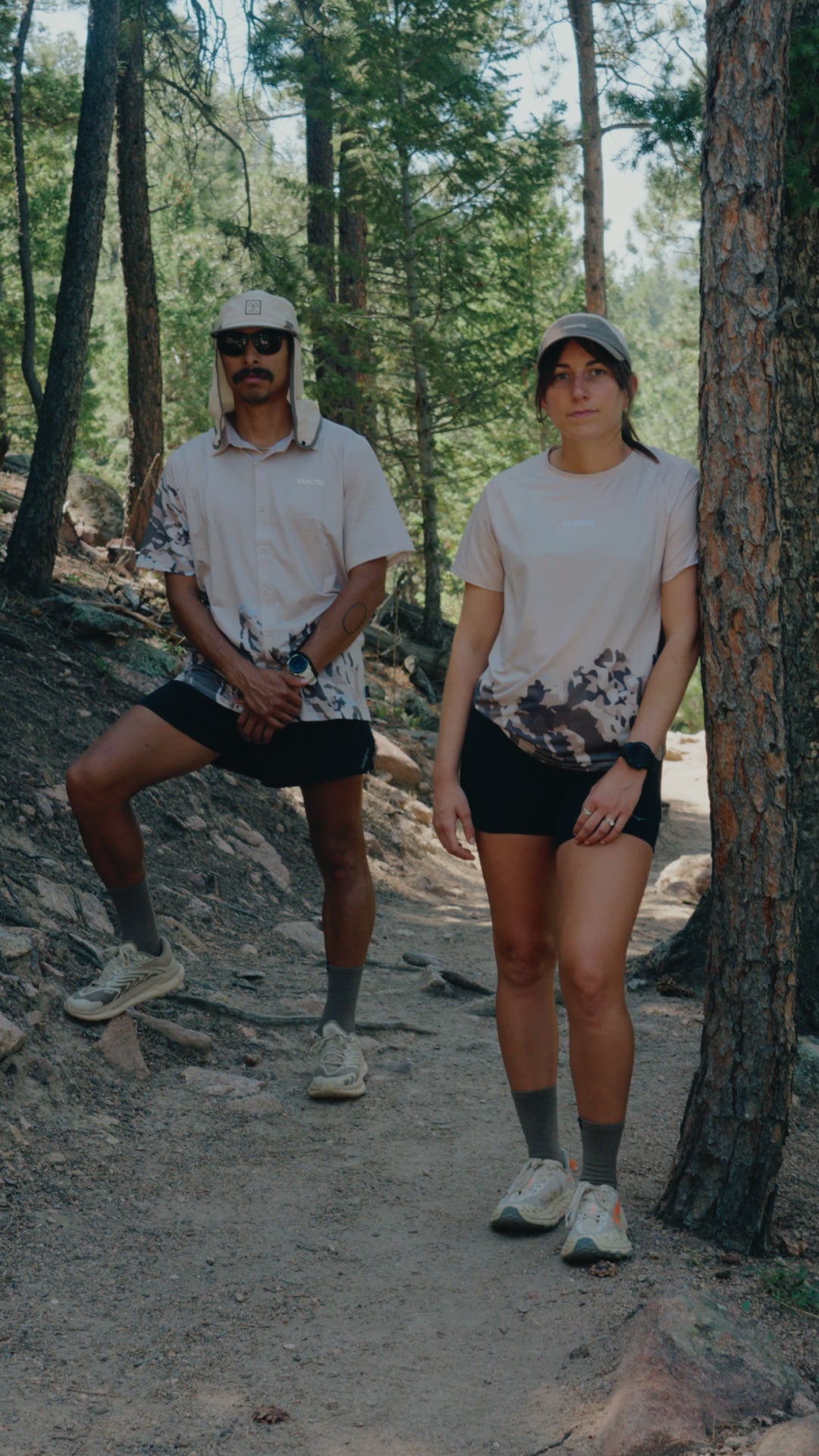 A man and a woman wearing matching beige and black outdoor activewear with a mountainous print, standing on a dirt trail in a pine forest.